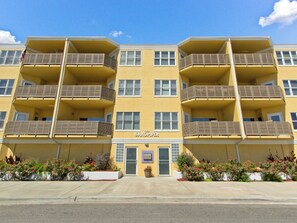 A yellow multi-story building with balconies, a sign reading "The Sandpiper," and a landscaped front with flowers. - Sandpiper 107 | Ocean Views, 20 Yards to Beach! (Tybee Island)
