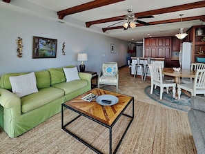 A living room with a green sofa, a wooden coffee table, a dining area with a wooden table and white chairs, and a kitchen with wooden cabinets and a refrigerator. - Ocean Song 321 | Pet Friendly, Oceanfront Condo! (Tybee Island)