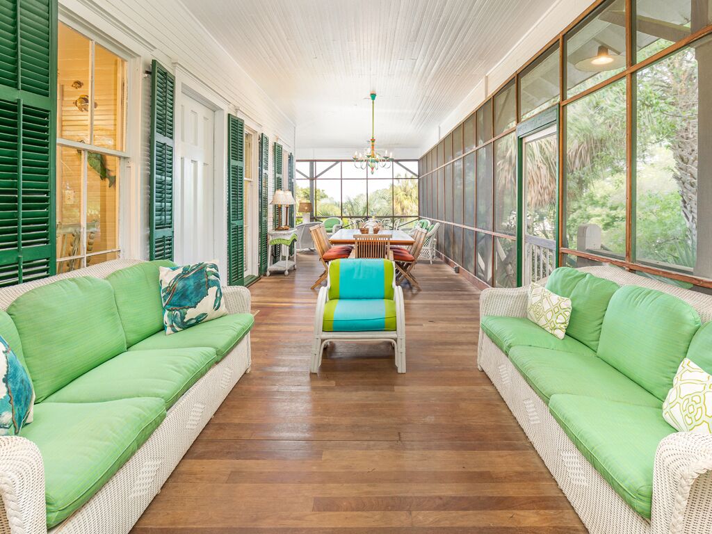 A spacious room with a wooden floor, green upholstered sofas, a dining table with chairs, and large windows offering a view of greenery.