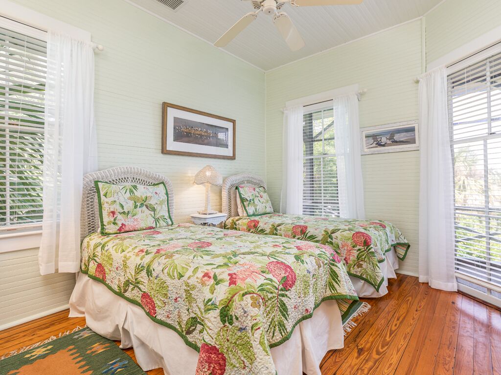 A bedroom with two beds, white curtains, wooden flooring, and ceiling fans.