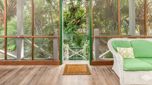 Une pièce dotée d'un plancher en bois, d'un tapis vert, d'un canapé en osier blanc agrémenté de coussins verts, et offrant une vue sur un jardin luxuriant grâce à de grandes fenêtres.