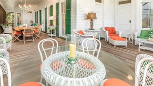 Une véranda lumineuse et spacieuse, meublée de meubles en osier, avec une table en verre décorée de bougies et offrant une vue sur la maison.