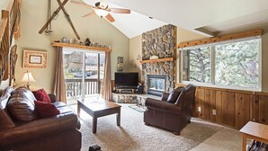 Ein gemütliches Wohnzimmer mit Kamin aus Stein, Ledersofas, Deckenventilator und einem großen Fenster mit Blick auf Bäume.