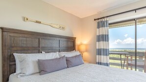 Une chambre à coucher avec un grand lit, une tête de lit en bois, une table de chevet, une lampe et une vue sur l’extérieur par une fenêtre ornée de rideaux.