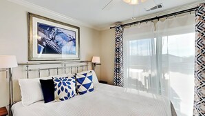 A hotel room with a large bed, a framed picture on the wall, a balcony with a view, and a ceiling fan. - Lighthouse Point 9C | Top Floor, Bay Views, Pools! (Tybee Island)