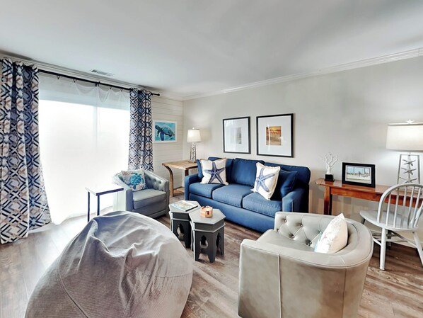 A modern living room with a blue sofa, a grey armchair, a round coffee table, and a white chair. There are two framed pictures on the wall, a lamp on a side table, and a window with patterned curtains.