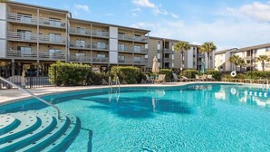 A swimming pool with a clear blue water feature, surrounded by a multi-story hotel building with balconies and a landscaped area with palm trees. - Lighthouse Point 9C | Top Floor, Bay Views, Pools! (Tybee Island)