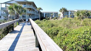 A wooden boardwalk leading to a beachfront hotel with multiple balconies and palm trees. - Lighthouse Point 9C | Top Floor, Bay Views, Pools! (Tybee Island)