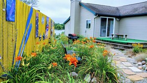 A house with a stone pathway leading to a green deck, surrounded by orange flowers and greenery. - Lakefront/Wifi /Coffee/ Stocked Cabin /Kayaks /Beach/Firepit hammocks /Pet (Harrison)
