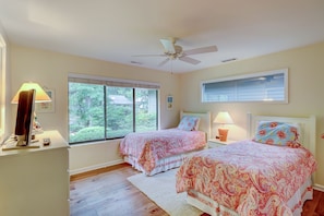 Une chambre avec deux lits, un ventilateur de plafond, un bureau avec une lampe et une fenêtre donnant sur un paysage verdoyant.