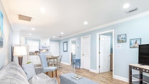 A living room with a grey sofa, a coffee table, and a TV stand. There is a kitchen in the background with white cabinets and a dining area with a table and chairs. - Dune B | Updated Ground Floor, Pet Friendly, 200 Yards to Beach (Tybee Island)