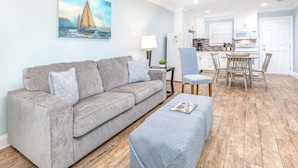 A living room with a grey sofa, a blue ottoman, and a painting of a sailboat. - Dune B | Updated Ground Floor, Pet Friendly, 200 Yards to Beach (Tybee Island)