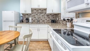 A modern kitchen with white cabinets, a granite countertop, a built-in stove, and a dining area with a wooden table and chairs. - Dune B | Updated Ground Floor, Pet Friendly, 200 Yards to Beach (Tybee Island)