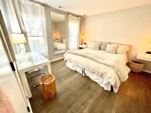 A bedroom with a bed, bedside table, lamp, mirror, and a small wooden stool.