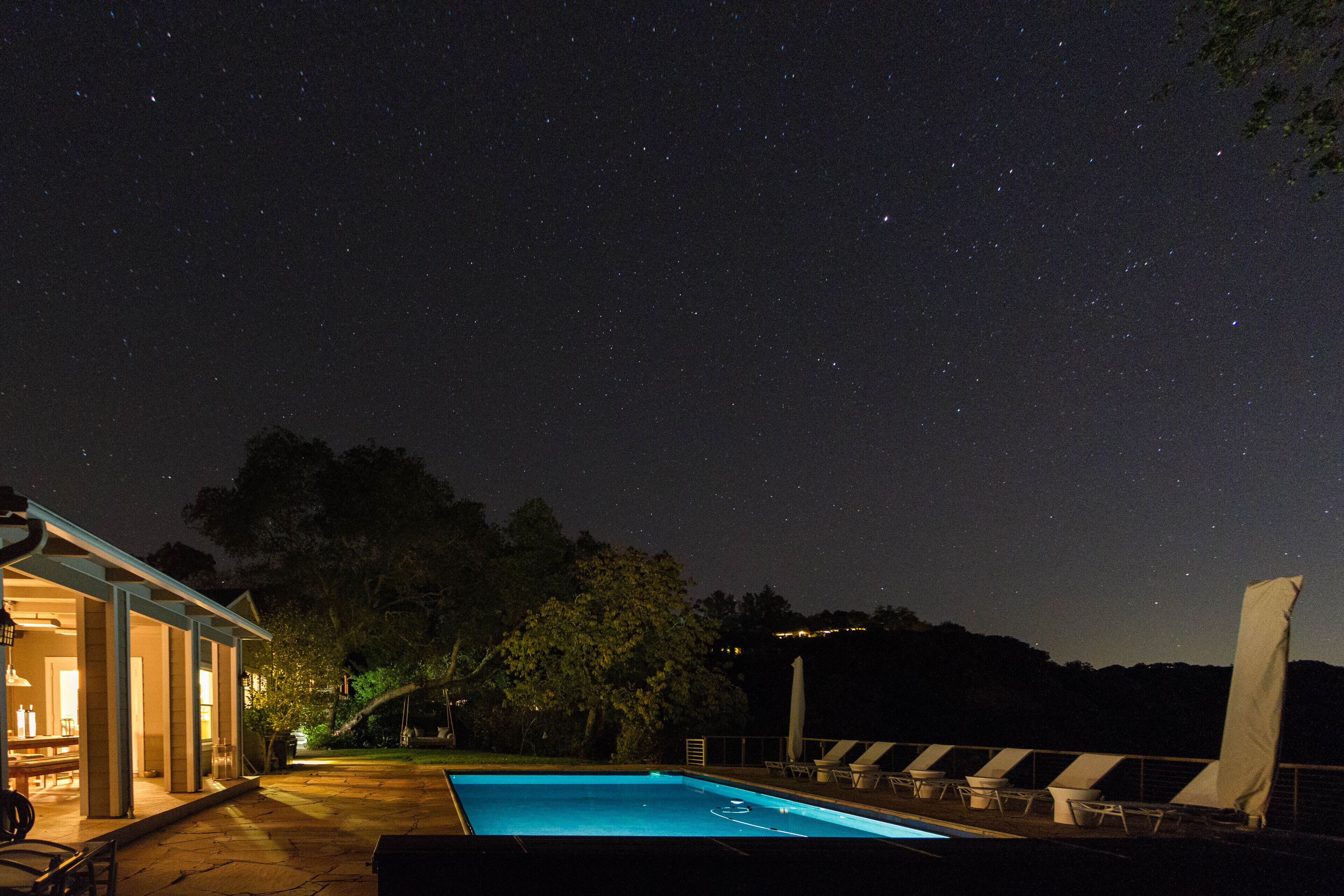 A swimming pool with lounge chairs and a covered area, surrounded by trees and a night sky with stars.