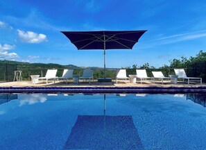 A pool with white lounge chairs and a large umbrella, surrounded by a clear blue sky and a hilly landscape.