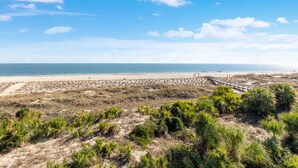 Ein Strand mit einer Holzpromenade, die zum Ufer führt, umgeben von Dünen und vereinzelter Vegetation.
