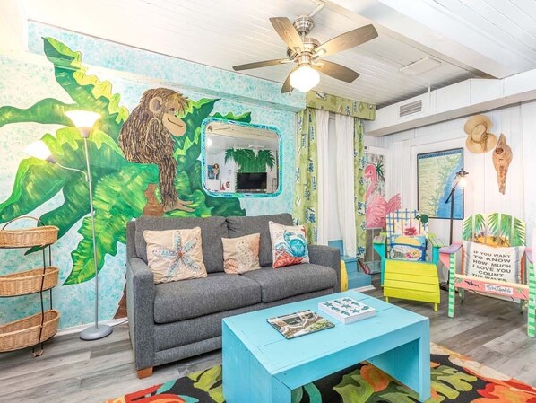 A room with a grey sofa, a blue coffee table, a ceiling fan, and a wall mural with a monkey and tropical leaves. - Tybreeza B | Apartment, Shared Pool, Near Beach! (Tybee Island)
