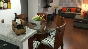 Un coin repas avec une table en verre, des chaises et une bouteille de vin posée sur la table. Le salon comprend un canapé et un téléviseur.