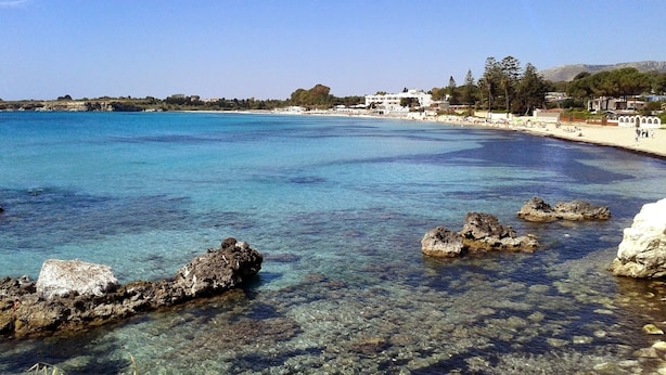 Una playa costera con aguas claras, formaciones rocosas y edificios al fondo.