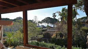 Una pérgola de madera con un banco acolchado y una mesa, con vista a un jardín exuberante lleno de plantas y una vista distante de casas.