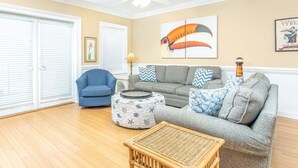 A living room with a blue armchair, grey sofa, and a wicker coffee table. There is a ceiling fan and a wall with a large toucan painting. - Private Balcony, Community Pool, 30 Yards to Beach (Tybee Island)