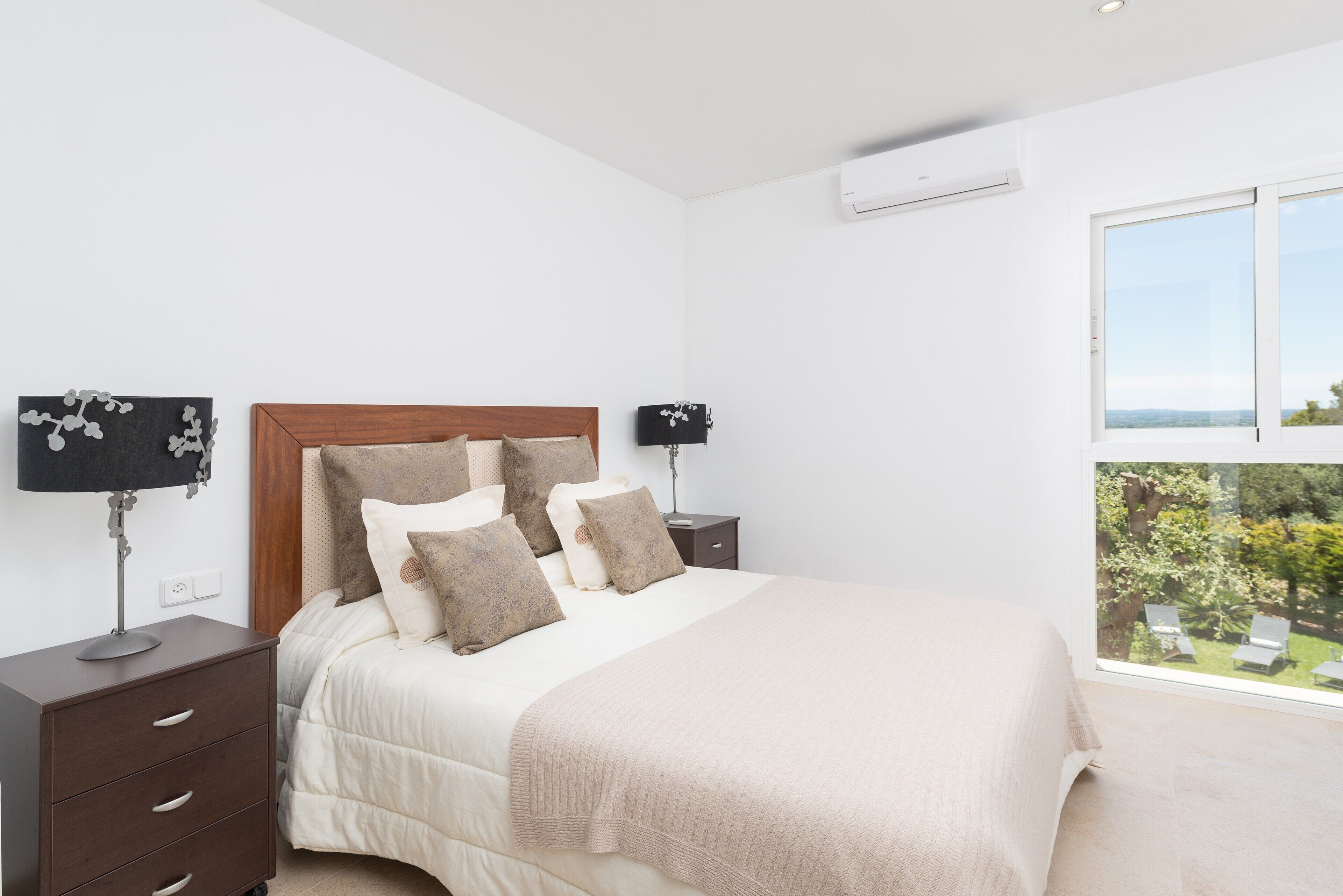 Une chambre dotée d’un grand lit, de deux tables de chevet, d’une commode, d’une fenêtre donnant sur un espace verdoyant et d’un appareil de climatisation.