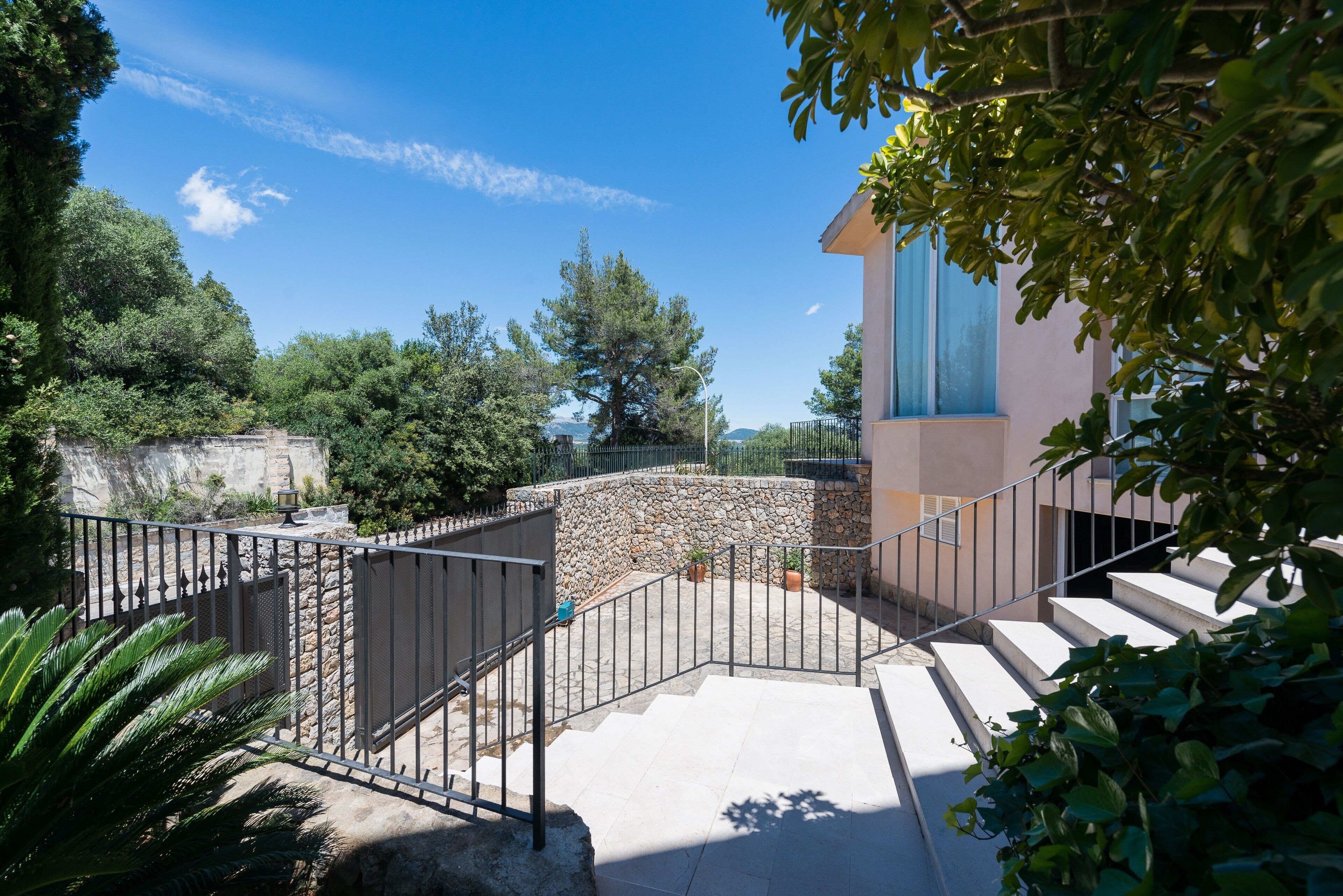 A stone staircase leading to a building with a blue window, surrounded by greenery and a clear sky.