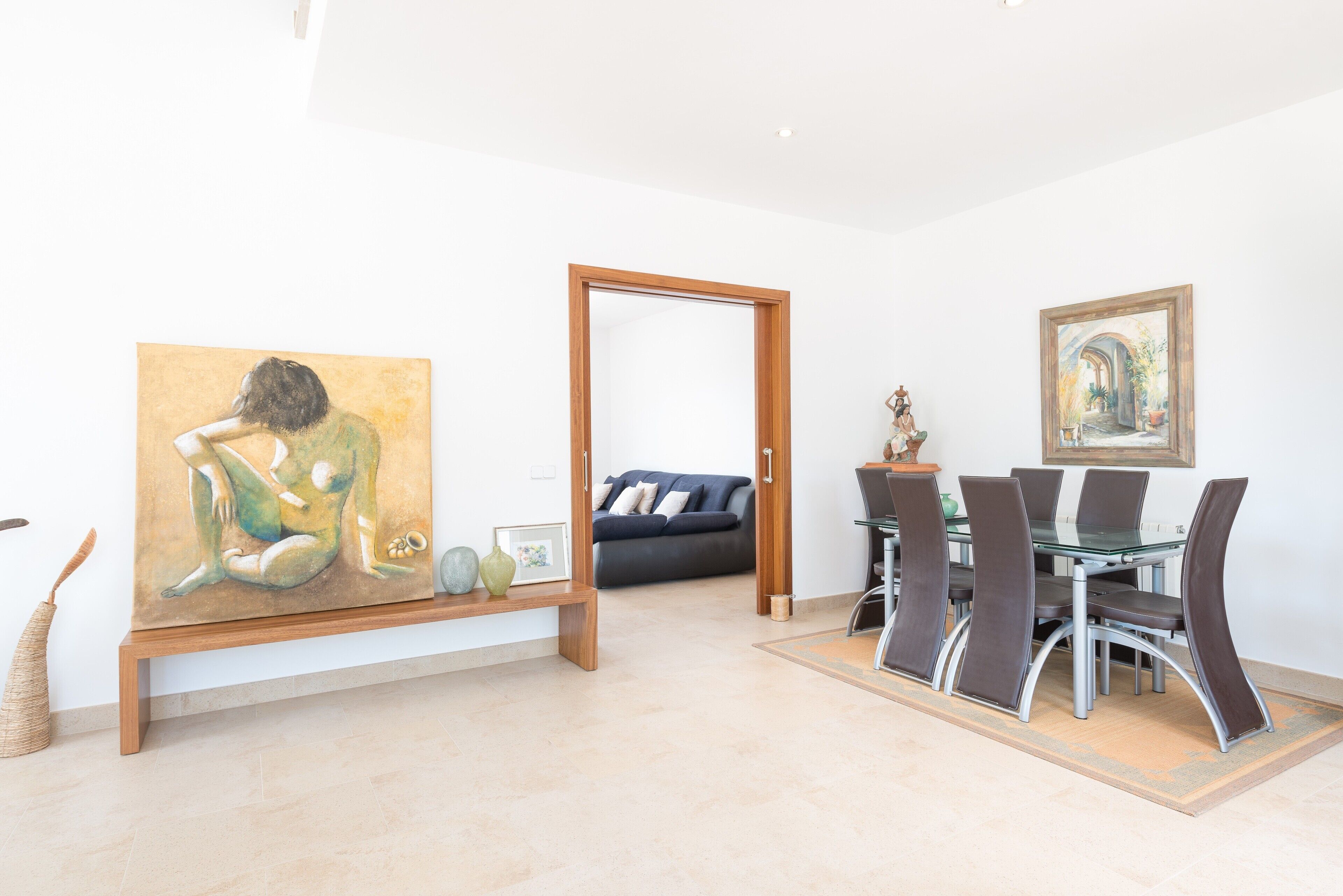 A modern living room with a dining area, featuring a sofa, a glass dining table with chairs, and artwork on the walls.