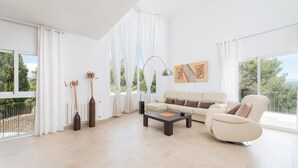 Un salon spacieux avec un canapé beige, une table basse et une lampe moderne. De grandes fenêtres, ornées de rideaux blancs, offrent une vue sur la verdure environnante.