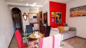 Un comedor con mesa y sillas de vidrio, una cocina al fondo y una pared roja decorada con obras de arte.