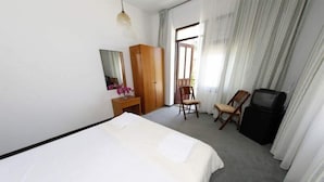 A hotel room with a bed, a wooden wardrobe, a desk with a mirror, a chair, a television, and a large window with curtains. - Mer Pansiyon (Fethiye)
