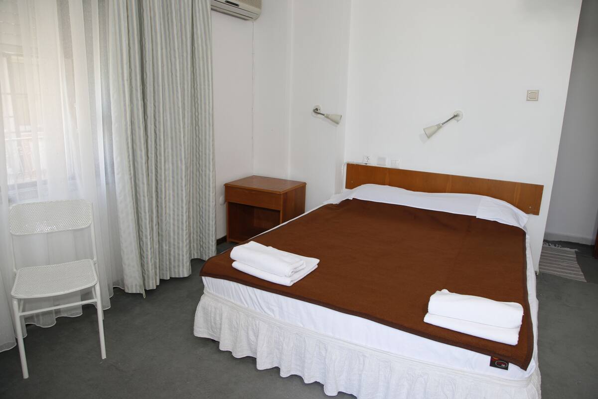 Una habitación de hotel con una cama individual, una mesita de noche de madera, una silla blanca y una ventana con cortinas de gasa.