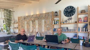 Dos hombres sentados en una mesa con computadoras portátiles, rodeados de estanterías y objetos decorativos.