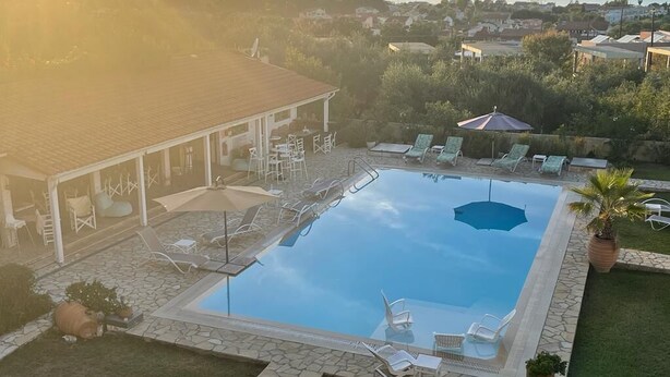 Una piscina al aire libre con sillones de descanso y sombrillas, rodeada por un patio de piedra y vista al mar y al pueblo al fondo.