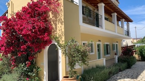 Un edificio de dos pisos con balcón, un camino de piedra y plantas de buganvilla.