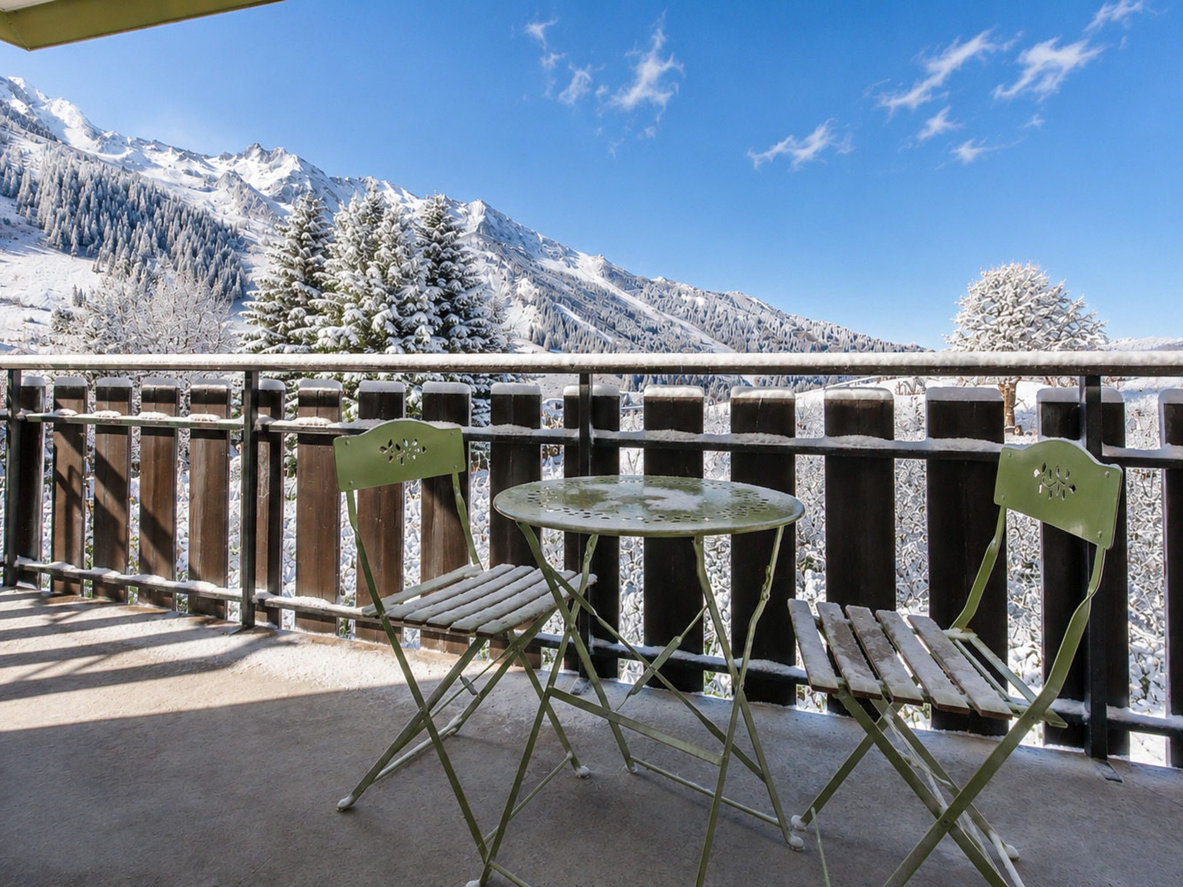 A balcony with a table and two chairs, overlooking a snow-covered mountain range.