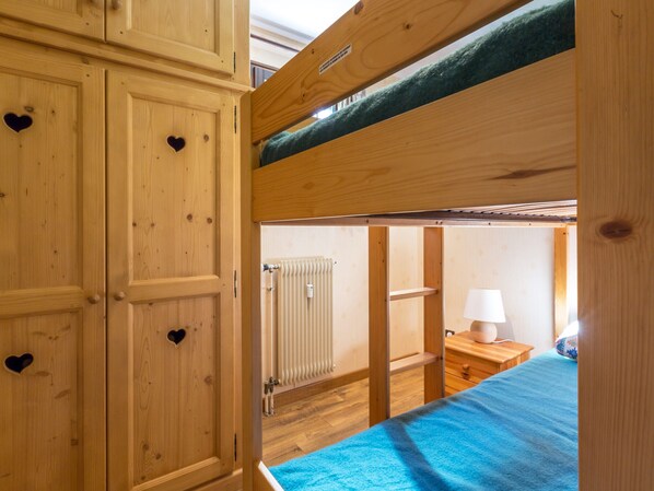 Une chambre avec un lit superposé en bois, une couverture bleue, une armoire en bois ornée de découpes en forme de cœur et une petite table de chevet avec une lampe.
