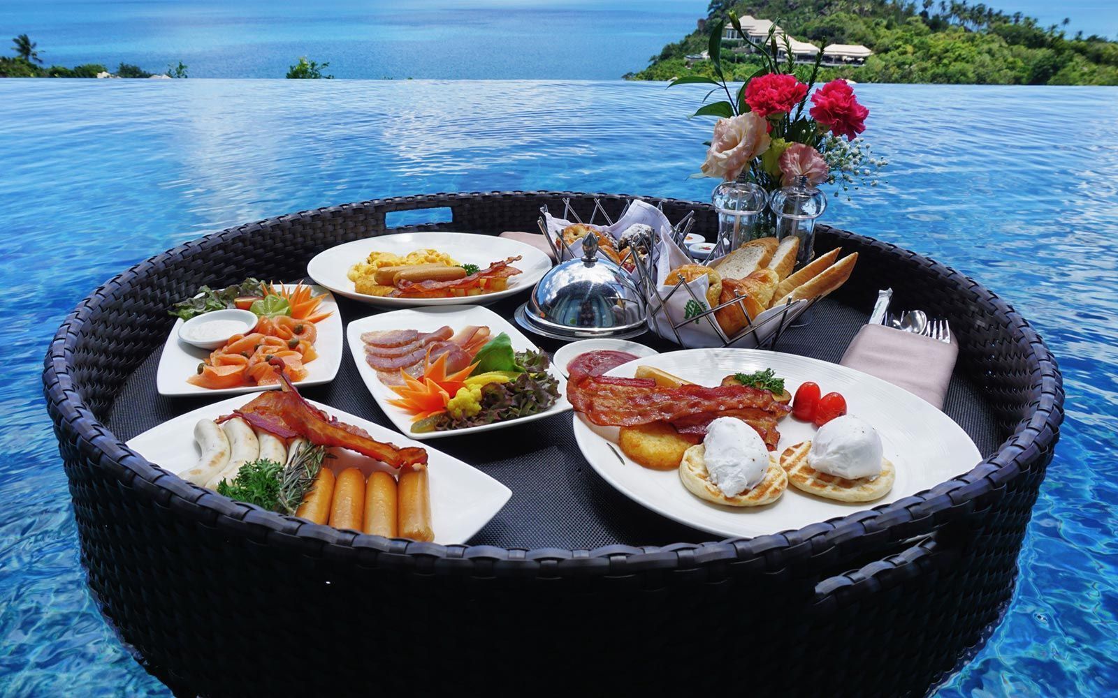 Un buffet de petit-déjeuner est dressé sur une table au bord de la piscine, avec divers plats, notamment des œufs, du bacon et des viennoiseries, le tout agrémenté d’un panier en osier et d’un vase rempli de fleurs.