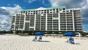Un grand hôtel à plusieurs étages, doté de balcons, situé sur une plage de sable et équipé de chaises longues et de parasols.