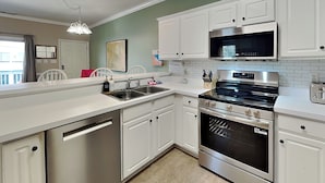 A modern kitchen with white cabinetry, stainless steel appliances, and a large island with a sink. - Tybee Sands 4A | Steps to Beach, Dining, Shops (Tybee Island)