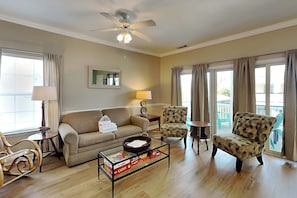 A living room with a ceiling fan, a sofa, two armchairs, a coffee table with board games, and a window with curtains. - Tybee Sands 4A | Steps to Beach, Dining, Shops (Tybee Island)