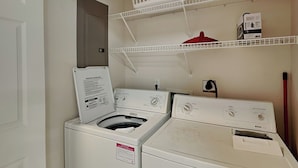 A laundry room with a front-loading washer and dryer, a white basket on a shelf, and a red pot on the dryer. - Tybee Sands 4A | Steps to Beach, Dining, Shops (Tybee Island)