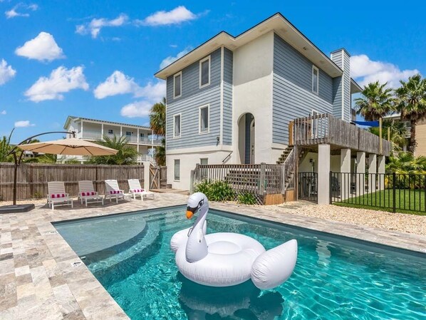 Ein Schwimmbecken mit einer Schwan-Luftmatratze, ein Haus mit Balkon und eine Holzterrasse.