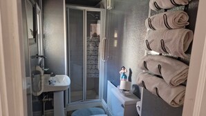 Un baño con cabina de ducha de vidrio, lavamanos y retrete. Hay toallas apiladas en un estante.