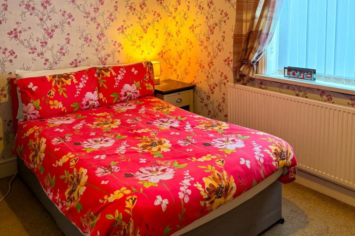 A bedroom with a floral-patterned bedspread, a bedside table with a lamp, and a window with curtains.