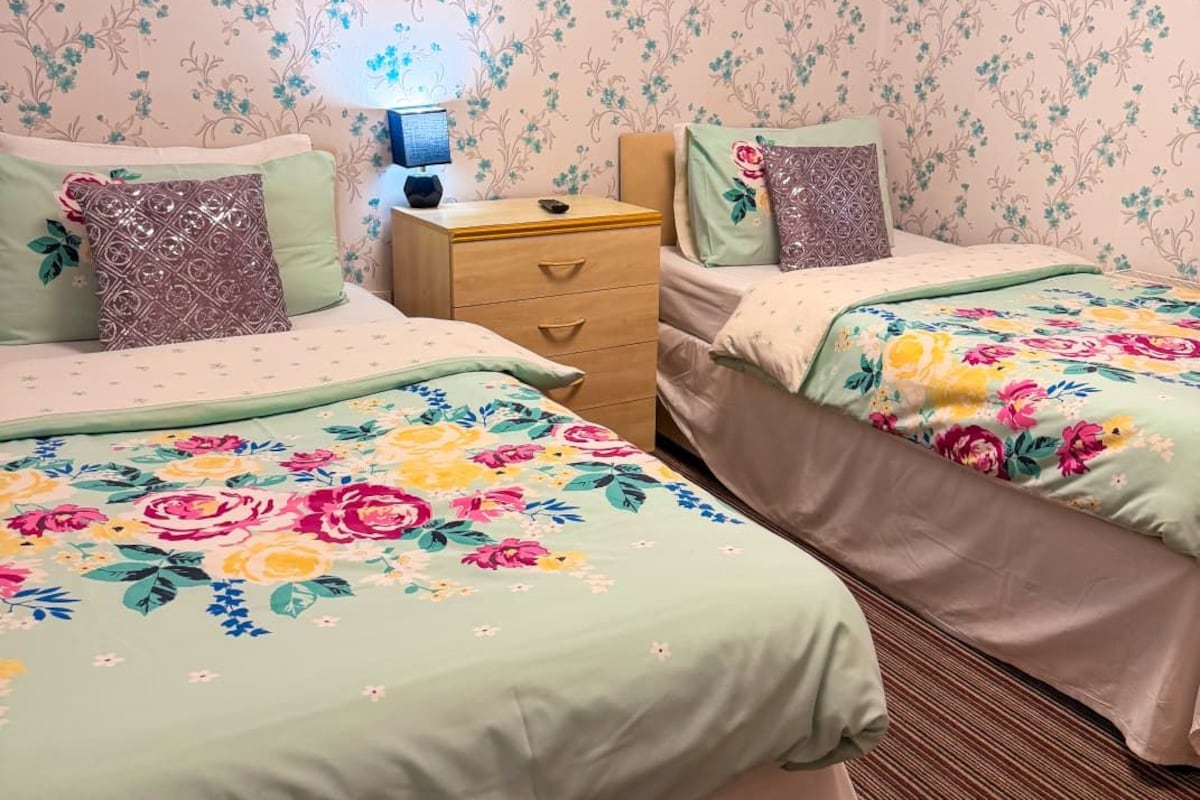 A hotel room with floral wallpaper, two beds with floral bedspreads, a nightstand with a lamp, and a wall decoration.