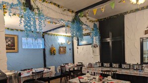 Un comedor con mesas dispuestas para comer, una zona de cocina con ollas y sartenes, y decoraciones colgantes azules y amarillas.