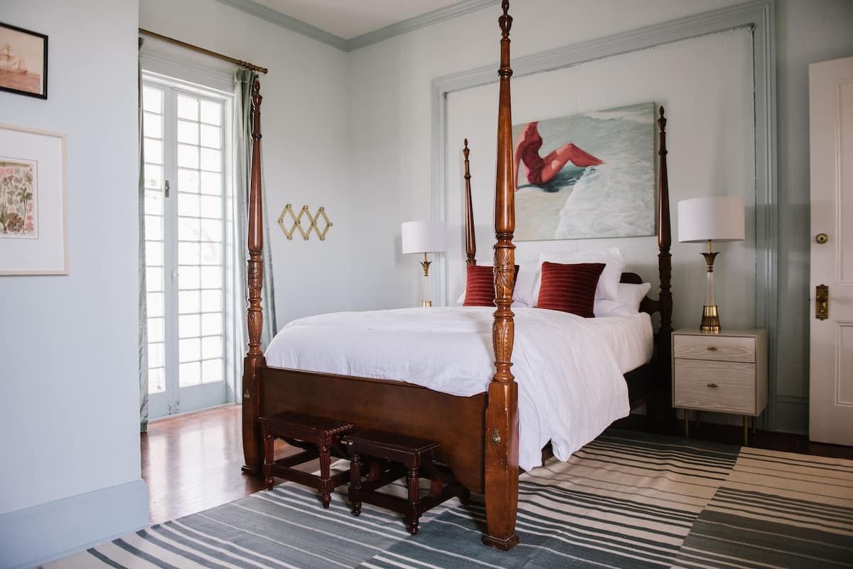 A four-poster bed with a wooden frame and white bedding, a nightstand with a lamp, and a painting above the bed.