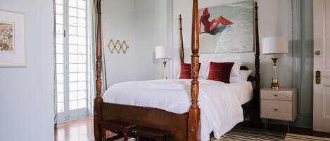 A four-poster bed with a wooden frame and white bedding, a nightstand with a lamp, and a painting above the bed.